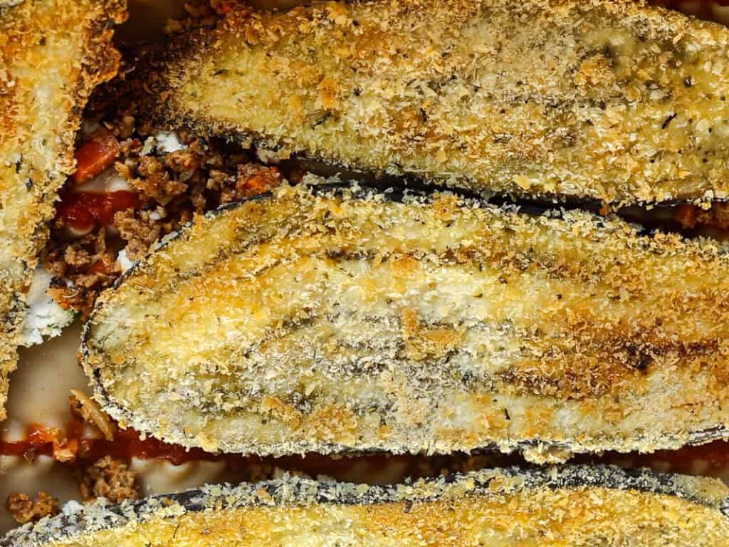 Close-up of baked eggplant slices coated in breadcrumbs, with a filling of minced vegan sausage and tomato sauce. The golden-brown crust indicates they are likely part of a baked dish, such as stuffed eggplants or eggplant parmesan.